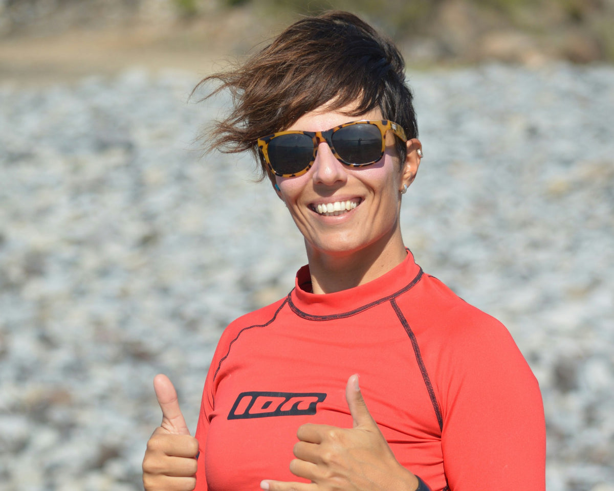 Woman in a red long-sleeve rash guard giving a thumbs up on a rocky beach